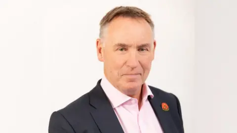 Head and shoulders image of Simon Bowen - he is wearing a dark blazer with a metal poppy badge and light pink shirt. He has short silver and brown hair.