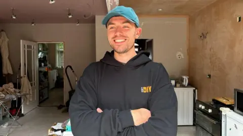 Scott Baggaley, a man wearing a black hoodie and a blue baseball cap, is stood with his arms folded in a room which is undergoing DIY.