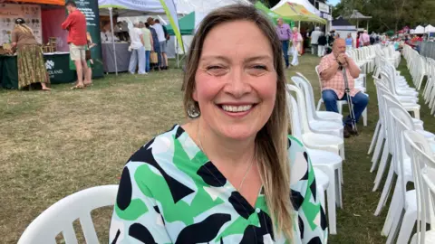 Claire Greenslade is sitting on a white plastic chair which is part of a row of white plastic chairs on a lawn. In the background are stands where people are looking at things that are displayed.