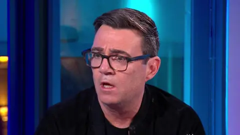 BBC Andy Burnham speaking on Newsnight. His face is animated in conversation and he is wearing dark-rimmed glasses and a black jumper.