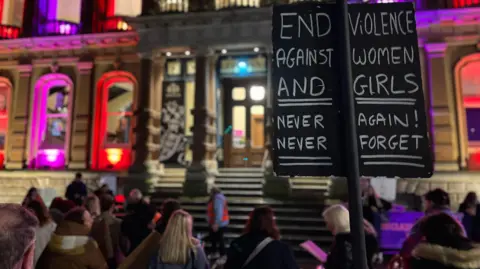 Jon Wright/BBC Suffolk A crowd outside Ipswich town hall at night. In the foreground is a placard which reads End violence against women and girls. Never again, never forget