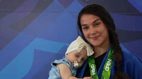 Bea Fernandes stands with her baby girl in her arms following a jiu-jitsu competition. She has a medal around her neck and has long dark hair that has been braided. Her baby wears a white hat with a bow on it and a blue dress with daisies on it. 