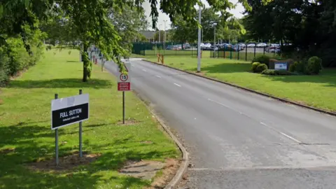 Google The entrance to HMP Full Sutton. A black and white sign stands on a neatly cut grass verge beside a roadway, with fencing and a car park in the background.