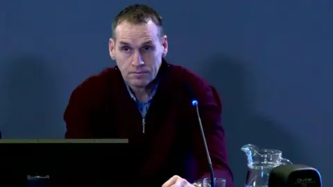 omagh bombing inquiry Ian Ferguson sits at a table with a computer and microphone in front of him. He  is wearing a maroon jumper with a blue shirt underneath. He has grey hair and brown eyes.