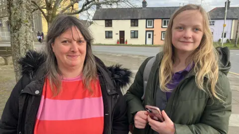 Sam Herbert, left, has long dark great hair and wears a black coat over a pink and red striped top. Stephanie, right, wears a green coat over a purple polo shirt and has long blonde hair. She is holding her phone and has a bag on one shoulder.