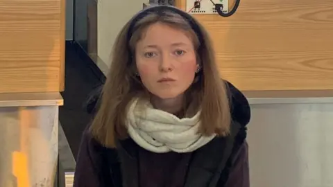 A woman with brown hair, dressed in black with a white scarf, sits in front of an airport check-in desk