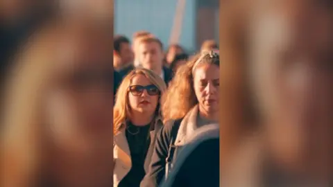 A crowd of people walk in the sunshine