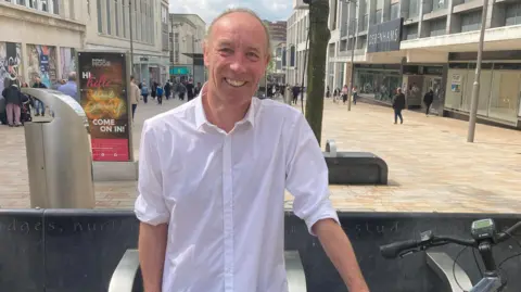 Photo shows a man with his hair scraped back, he is smiling and wearing a white shirt with the sleeves rolled up. He is stood on a city centre street with shops and pedestrians in the background