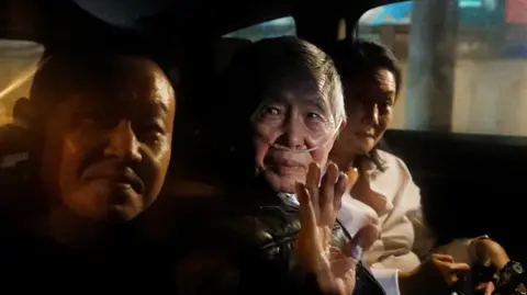 Getty Images Former Peruvian president Alberto Fujimori sits in the middle seat at the back of a vehicle, waving, as he leaves Lima's Barbadillo prison. His daughter Keikio is sat next to him wearing white and another male is to Fujimori's right. Fujimori has a supply of oxygen running through his nostrils.