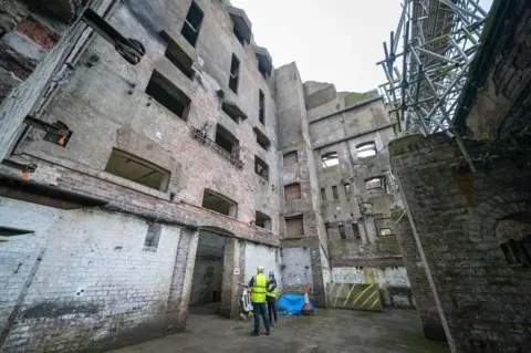 PA Interior courtyard of a large derelict industrial building with exposed brickwork, empty window openings and weathered concrete walls. Two construction workers wearing high-visibility clothing and hard hats stand on the ground floor inspecting the site, while extensive metal scaffolding rises along one side of the structure. The scene is lit by daylight from above, highlighting the building's deteriorated condition and multiple storeys.