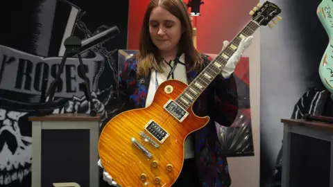 PA Images A woman is holding a guitar while wearing white gloves with Guns N' Roses posters behind her