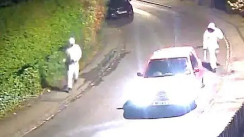 North Yorkshire Police A CCTV image shows two men wearing white hazmat suits walking down a residential street with machetes. A car with headlights on is parked on the street.