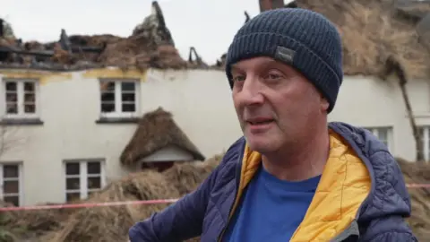 BBC A man who is dressed in a blue jacket and blue woollen hat is standing in front of a cottage with the thatched roof severely damaged by fire.