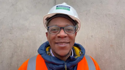 A head shot of a Severn Trent worker in a white hard hat and an orange hi-vis jacket. The man is smiling and standing in front of a wall.