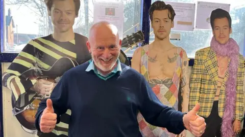 Zoe Toase / BBC Graham Blake sonríe y señala sus pulgares hacia arriba de tres cortes de cartón de Harry Styles en varios atuendos llamativos.