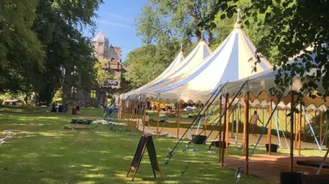 Outside Arts Four white marquees with wooden poles stand on a grassy plain. Huge green trees tower over the canvas. Behind them in the distance is a large stone building.