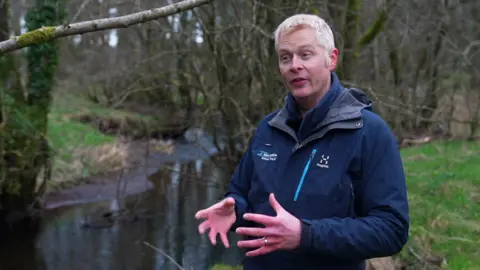 BBC WCRT assistant director Luke Bryant stands in front of river talking to camera. He is wearing blue jacket. 