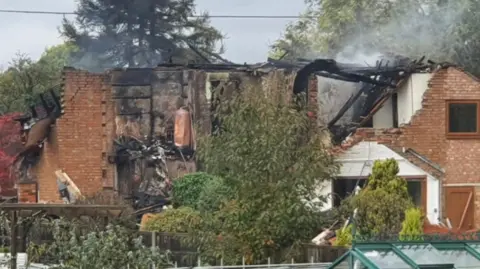 The photo shows the destroyed remains of a house with smoke coming from the roof.