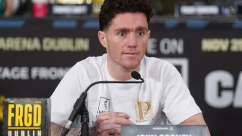 The late John Cooney pictured at a press conference.   He has short, dark, curly hair and blue eyes and is wearing a white t-shirt.   He is sitting in front of a microphone. 