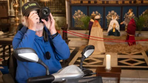 Lee Pullen A man in a wheelchair, his face covered by a large camera lens, with a scene of the nativity in the cathedral behind him