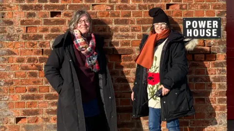 Dr Jane Watt, left, and Dr Susan Barnet, right, standing together outside a brick building on Orford Ness. A sign with the words Power House can be seen over the shoulder of Dr Susan Barnet. Shingle is visible to the left of the building, stretching into the distance