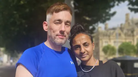A man with short, greying ginger hair and a beard in a blue sports top. A woman with long black hair in a bun, a beaded necklace and black top. They stand together with one arm around the other.