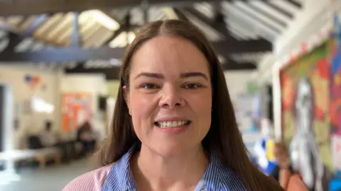 A woman with long brown hair is looking at the camera. She is wearing a red and blue striped shirt.