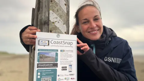 A lady with long brown hair tied back in a ponytail wearing a navy coat with GSNI written on it. She is standing beside a 'CoastSnap' poster which is pinned to a wooden post.