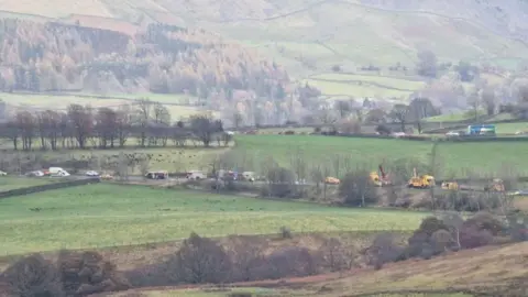 A Lake District scene with hills and fields and, in the middle of the image, there is a road containing a number of emergency vehicles