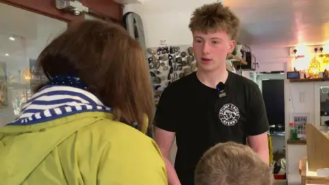 BBC/Seb Cheer Kayden, a teenager, speaks to a mother and son in a shop setting. He is wearing a T-shirt with "Stump Cross Caverns" written on it.
