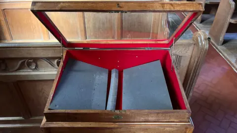 Martin Giles/BBC An empty wooden and glass display case in St Mary's. The glass lid is open and there is foam padding on the bottom of the case. The case is near a row of wooden pews.