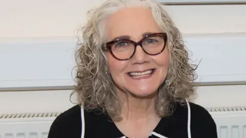 Mary with curly, shoulder-length grey hair wearing a dark top with white trim, seated indoors against a light-coloured wall and radiator.