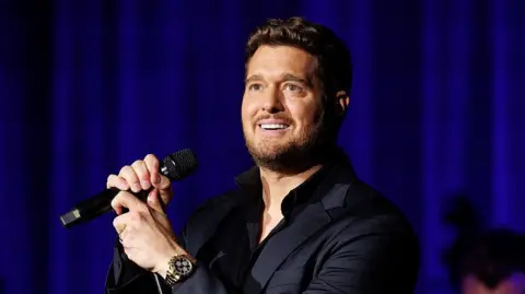 Michael Bublé. He has dark brown hair and beard, wearing a dark suit and watch. He is holding a microphone. 