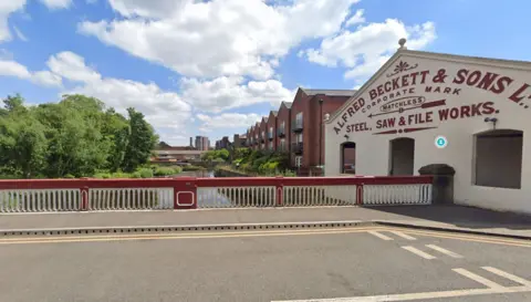 Ball Street Bridge above Kelham Island Weir: a red and white road bridge over a river, with buildings to the right and trees on the left. There is a weir in the distance, and tower blocks can be seen in the distance. 