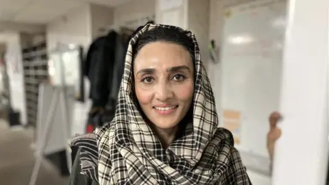 A woman in a black and white checked headscarf smiles at the camera. The background behind is blurred but she is standing in a white room with various objects in it.