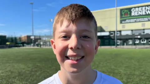 A boy wearing a white football top. He has brown hair. He is smiling. Behind him is the LNER Stadium.