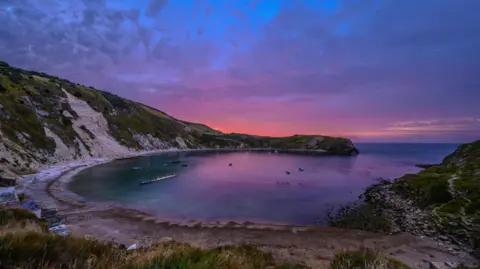 Richard Murgatroyd A sunrise over Lulworth Cove in Dorset. The sky is illuminated pink as the sun comes over the horizon behind the cliffs. Lulworth Cover is a semi-circular bay with shallow cliffs behind. There are some small boats in the bay and a grey beach. The cliffs have grass on them. 