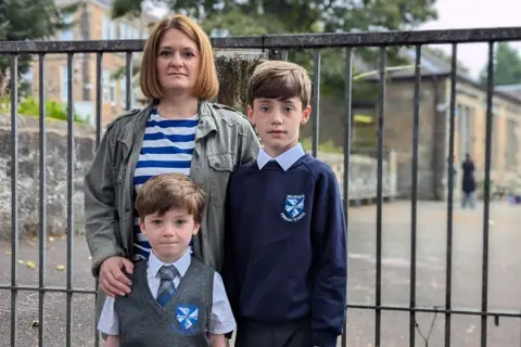 Julie, dressed in a striped top and green jacket, stands at the school gate with her arms around her songs in their primary school uniform.