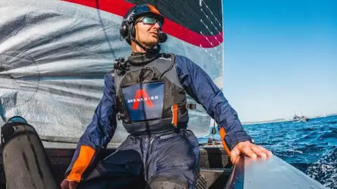 Giles Scott wearing a blue helmet with headset and blue waterproofs, on a yacht with a blue, grey and white sail behind him. The sea is visible to his left, and there is a boat in the distance.