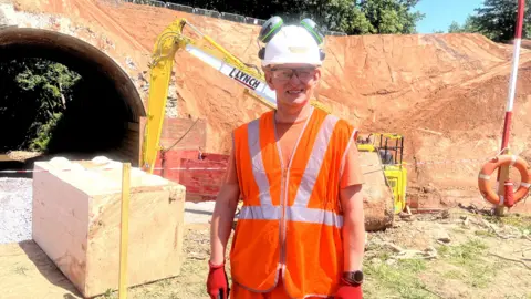 A man is wearing an orange hi-vis jacket with red gloves and a white helmet with grey headphones. In the background is a concrete block and a yellow digger. There is an arch bridge, the embankment is brown
