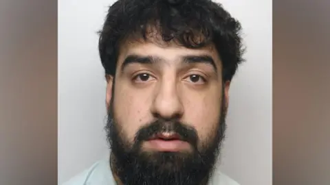 Custody photograph showing a person with dark hair and a full beard facing the camera against a plain light-coloured background.