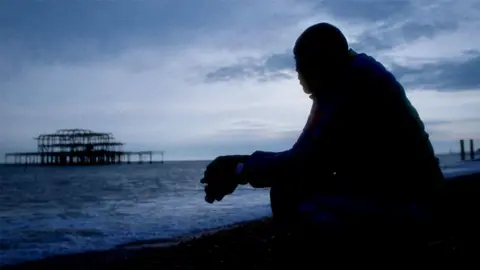 Paul A man sits on a beach at dusk, staring at the charred ruins of a sea pier.