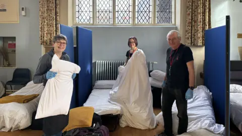 HopeWorks Three people making beds in a room, that has a large window with curtains either side. There are a number of beds in the room, and white sheets on them. A man, to the right, has on a black T-shirt and trousers and is wearing blue gloves. One woman is holding a pillow, with short hair and another woman holds a white duvet. 