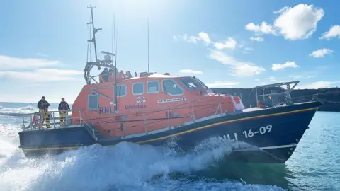 An RNLI lifeboat at see. A side on view of the blue and orange boat with people stood on the deck as it moves through the water. 