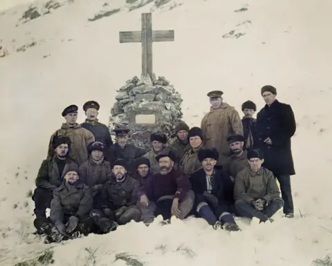 Autocromos originais de GH Wilkins, Coleção Rowett-Chojecki Um grupo de 16 homens da tripulação do navio de exploração Quest, de pé e sentados em frente a uma cruz memorial a Sir Ernest Shackleton