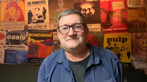 Jasmine lowe / BBC Paul Heaton who has short dark hair and is wearing glasses and a blue jacket and black T-shirt. he is standing in front of a wall of band posters at the New Adelphi Club