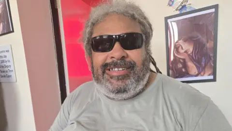 A man with greying dreadlocks and beard, and wearing sunglasses, smiles at the camera. He is sitting in a room with pictures on the white wall and a pink door behind him