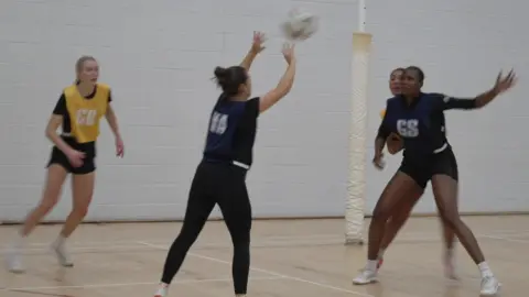 Four women training in a netball game, two have navy vests and two have yellow vests. 