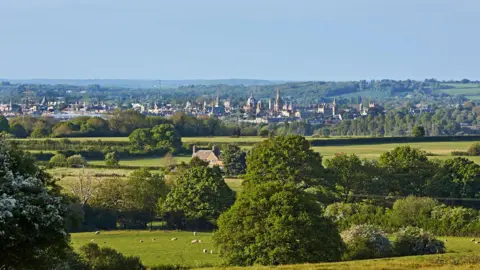 Getty Images A sunny summer day in Oxfordshire. Oxford city is in the distance with its spires visible. There are trees and green fields in the foreground. 