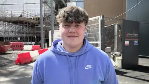 Elliot, in a blue hoodie, smiles at the camera and stands in front of a construction site